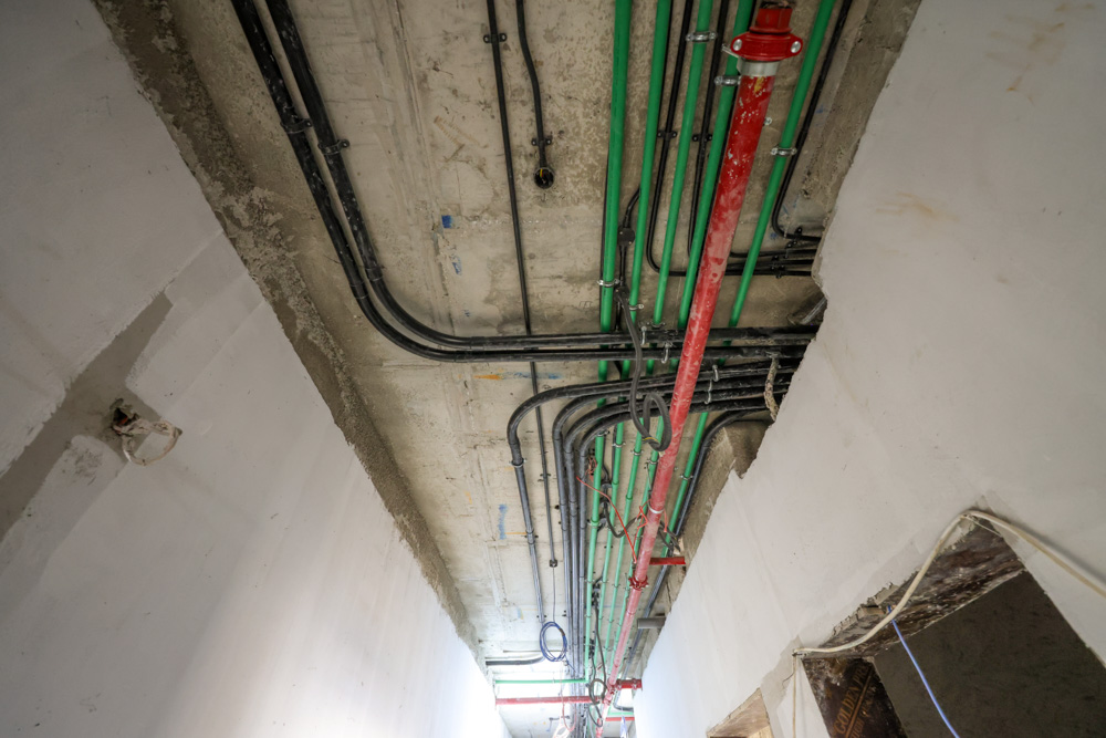Beach Walk Iii By Imtiaz Exposed pipes and electrical wiring on a concrete ceiling in an unfinished corridor with white walls.