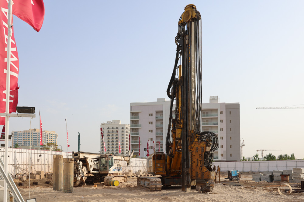 Cove Grand By Imtiaz A large drilling machine on a construction site with buildings and flags in the background under a clear sky.