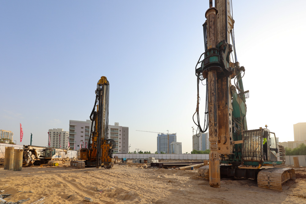 Cove Grand By Imtiaz Construction site with two large drilling machines operating, surrounded by buildings and sand.