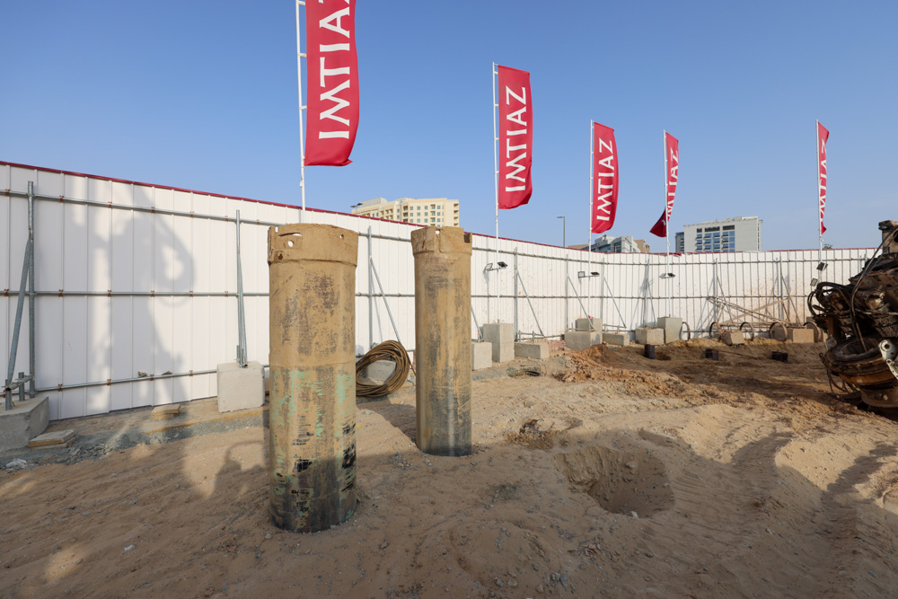 Cove Grand By Imtiaz Construction site with two large rounded pillars, flags, and equipment amid sandy terrain and a backdrop of buildings.