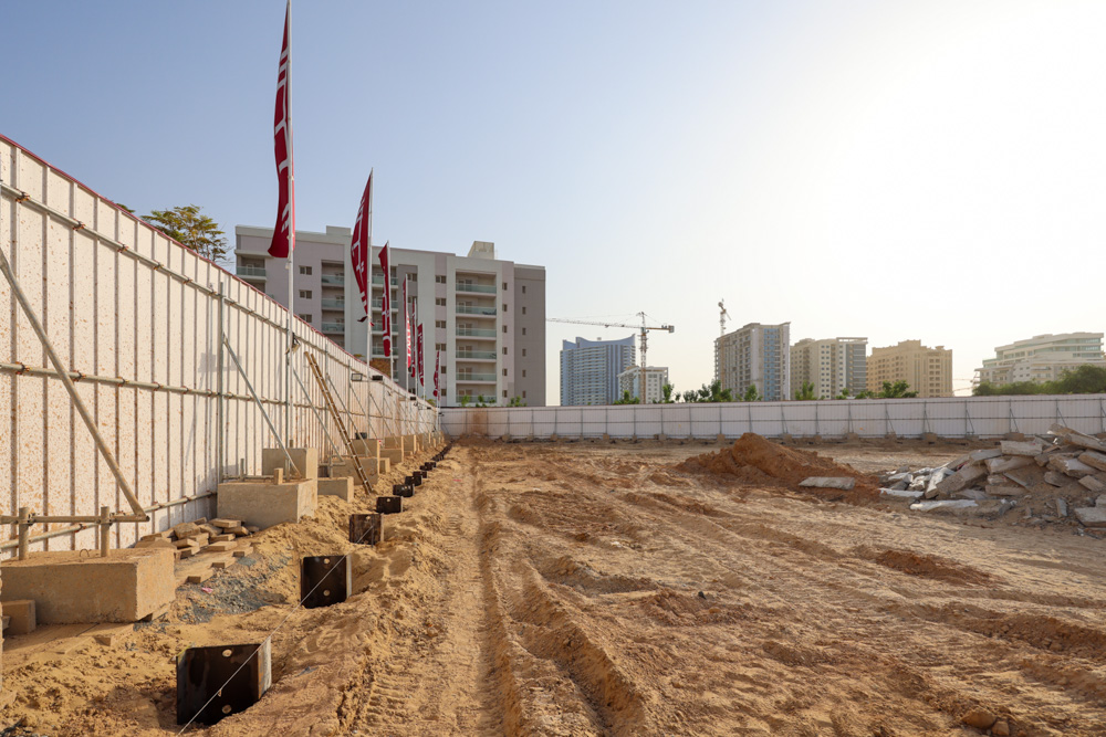 Cove Grand By Imtiaz Construction site with sandy ground, walls, flags, and buildings in the background under a clear sky.