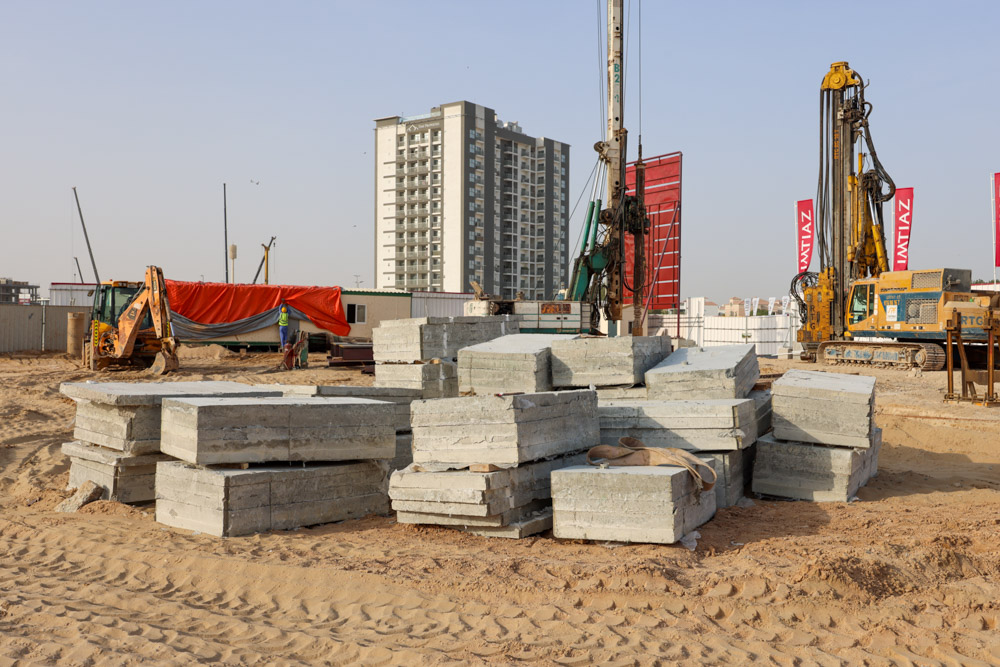 Cove Grand By Imtiaz Construction site with stacked concrete blocks, machinery, and a tall building in the background. Dusty ground visible.