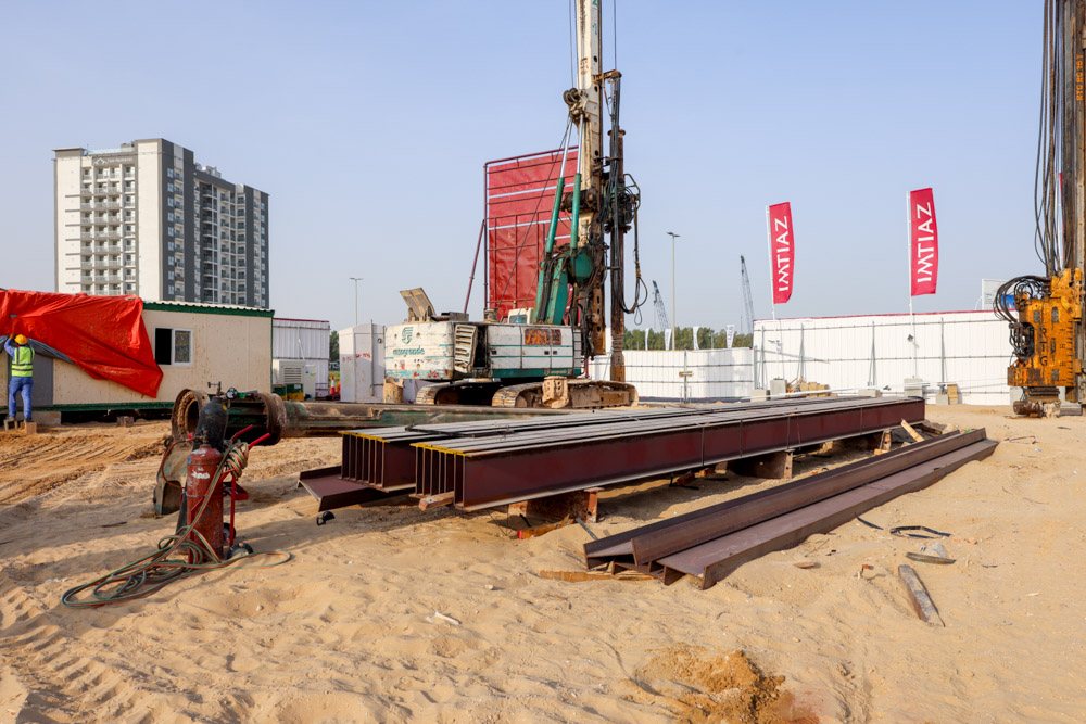 Cove Grand By Imtiaz Construction site with heavy machinery, steel beams, and a worker near a portable office against a backdrop of buildings.
