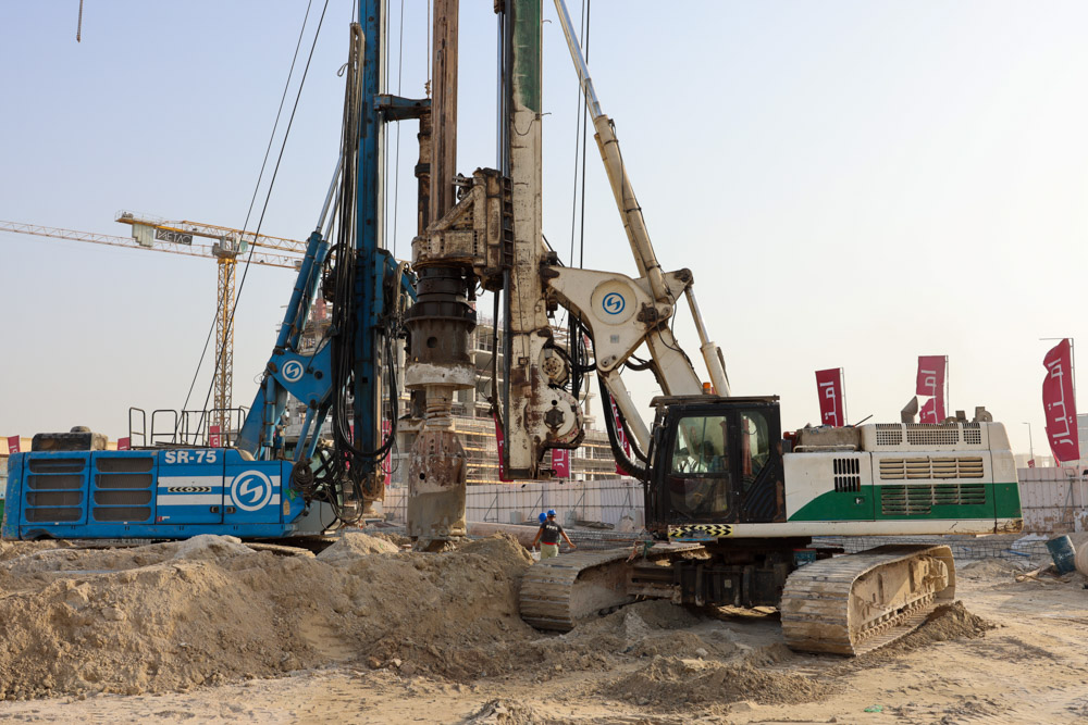 Cotier House 2 By Imtiaz A construction site featuring a drilling rig and machinery on sandy terrain, with cranes and workers in the background.