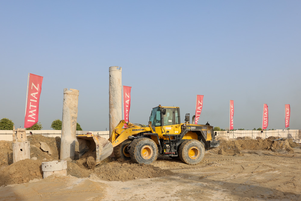 Cotier House 2 By Imtiaz A construction site with a Komatsu loader, concrete pillars, and flags displaying the name "IMTIAZ" in the background.