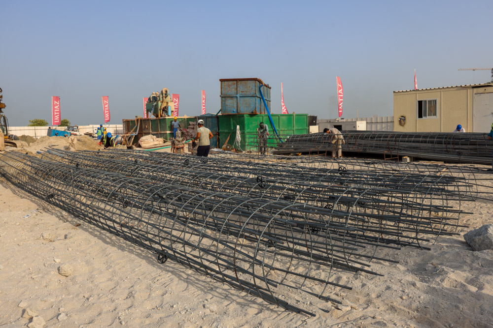 Cotier House 2 By Imtiaz Construction site featuring piles of rebar, workers, machinery, and storage containers under a clear sky.