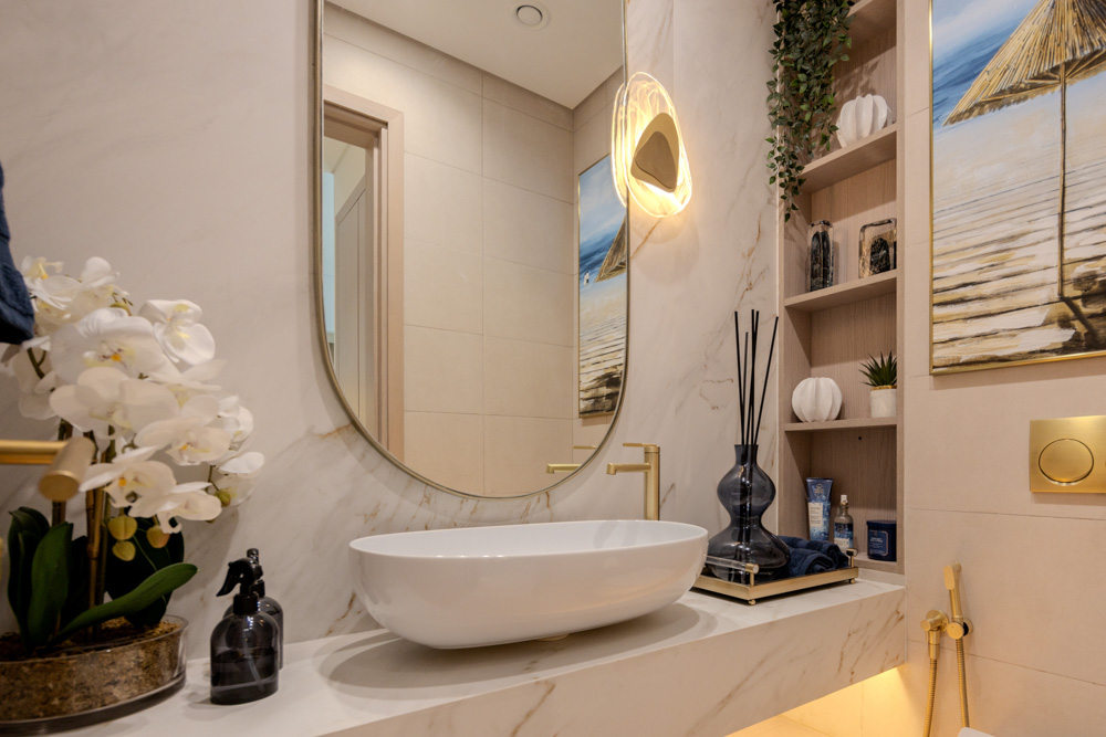Beach Walk Residence A modern bathroom featuring a round mirror, a white basin, decorative plants, and a wall painting of a beach scene.