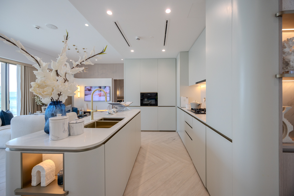 Beach Walk Residence Modern kitchen with white cabinets, gold accents, a large island, and decorative flowers in a blue vase.