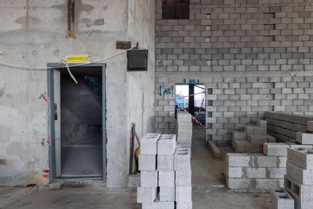 Beach Walk Iii By Imtiaz Interior view of a construction site with concrete walls, a staircase door, and stacks of stone blocks.