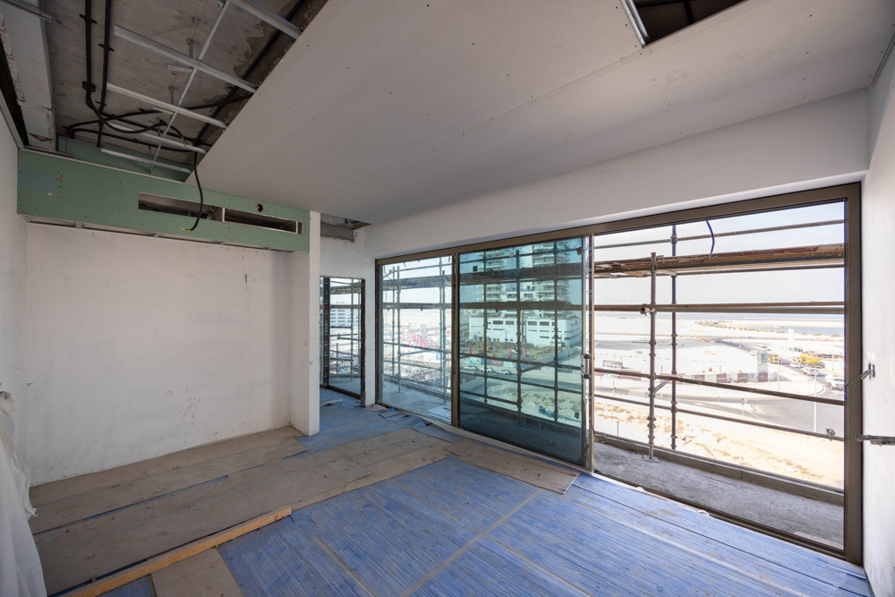 Beach Walk Iii By Imtiaz Empty room under construction featuring large windows and a view of a seaside area, showcasing scaffolding.