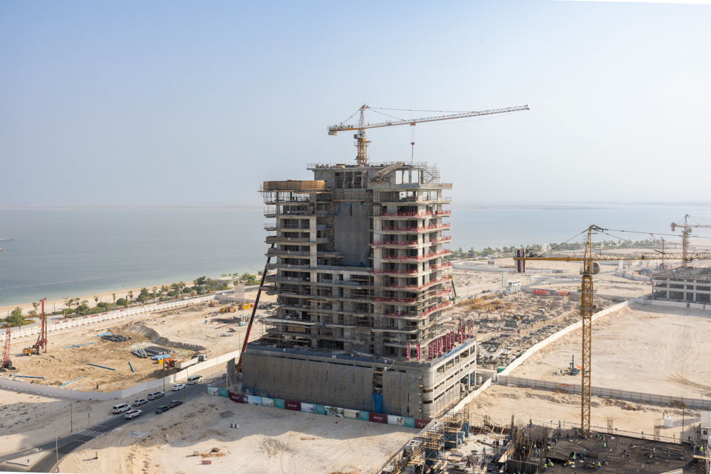 Beach Walk Iii By Imtiaz A tall building under construction with cranes, surrounded by a sandy area and a view of the water in the distance.