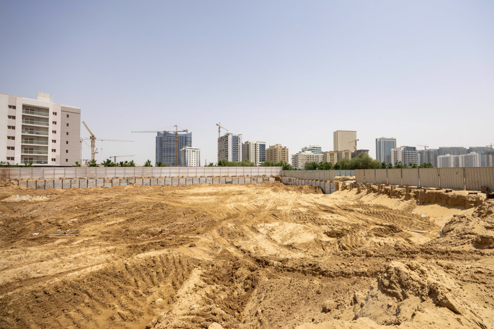 Cove Grand By Imtiaz A construction site with sandy terrain and nearby modern buildings and cranes under a clear blue sky.