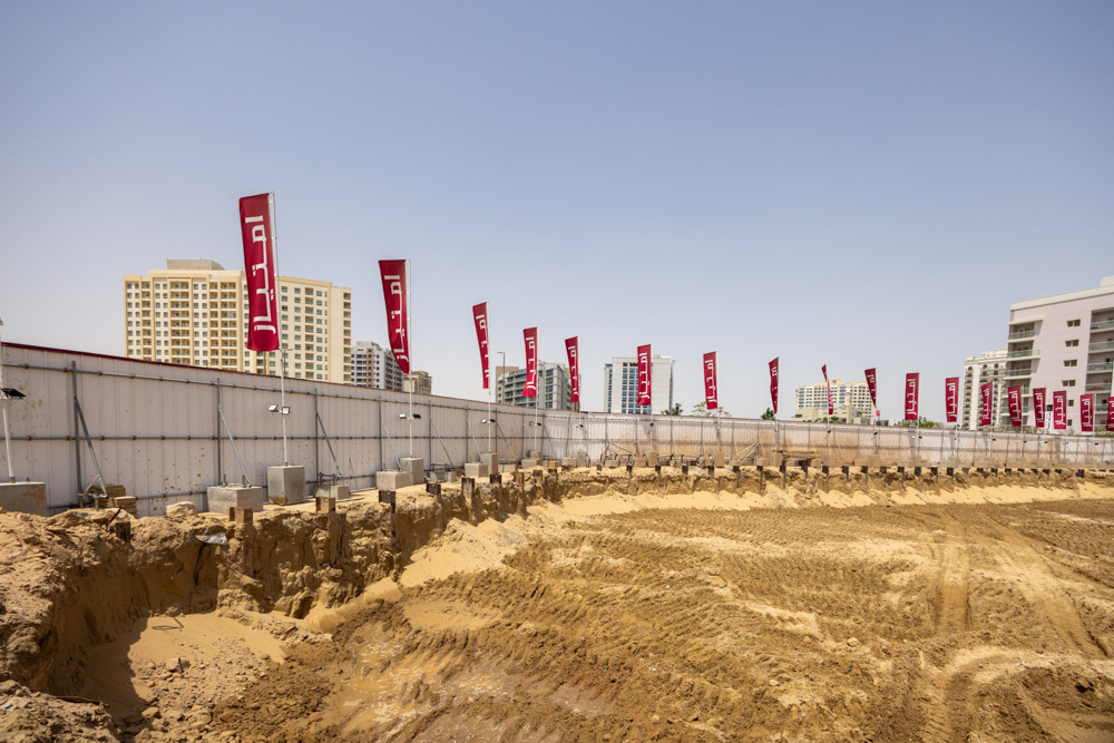 Cove Grand By Imtiaz Construction site with a fenced area, sand, and tall buildings in the background, featuring red banners.