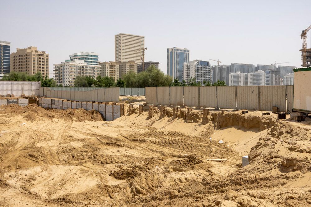 Cove Grand By Imtiaz A construction site with sand, soil, and a fenced area, surrounded by tall buildings in the background.