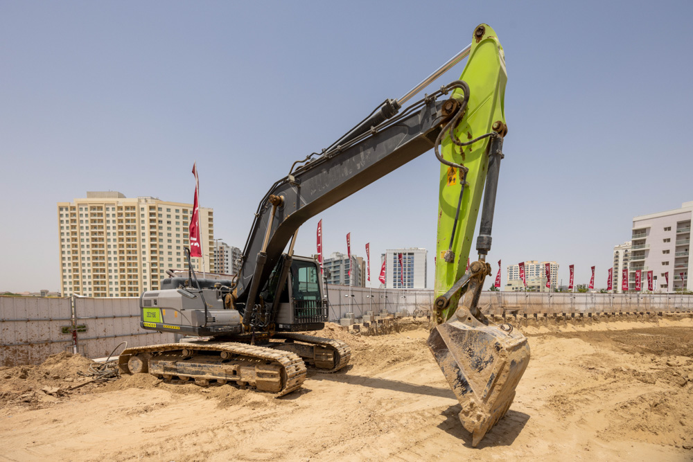Cove Grand By Imtiaz A large excavator with a green arm is positioned on a construction site with buildings and flags in the background.