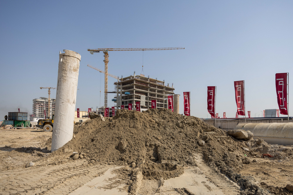 Cotier House 2 By Imtiaz Construction site with a large pile of dirt, concrete pillars, cranes, and red banners under a clear blue sky.