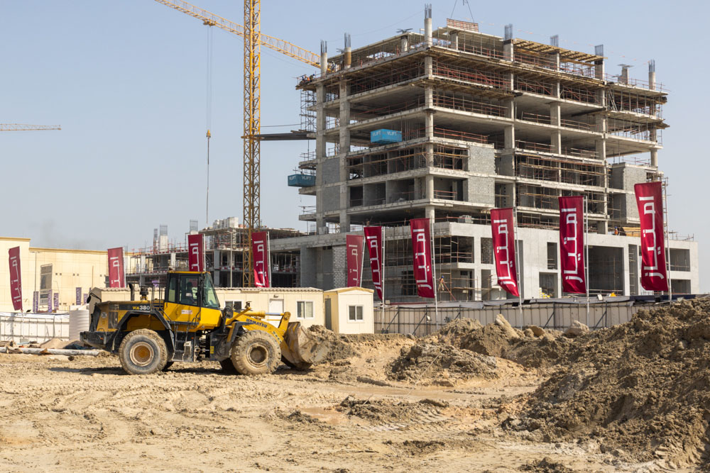 Cotier House 2 By Imtiaz Construction site featuring a partially built structure, a yellow bulldozer, and several banners in the foreground.