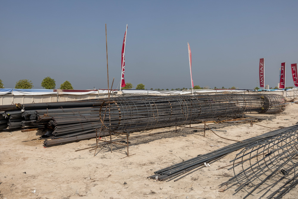 Cotier House 2 By Imtiaz A construction site with stacked steel rebar and mesh, surrounded by tents and flags under a clear blue sky.