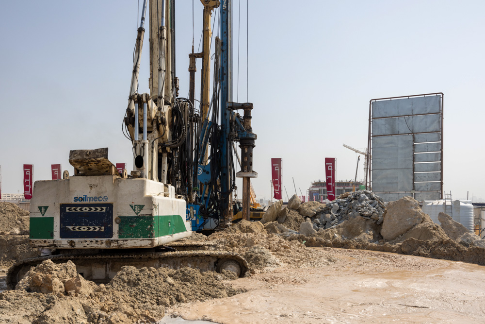 Cotier House 2 By Imtiaz A drilling machine at a construction site amidst large piles of rubble and construction materials under a clear sky.