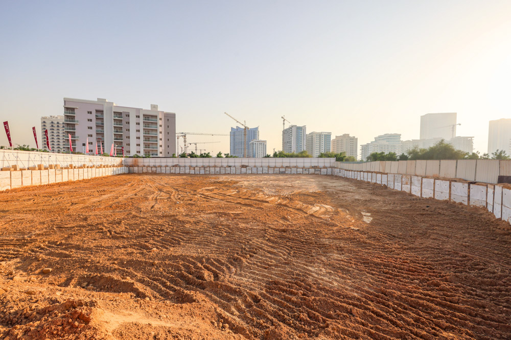 Cove Grand By Imtiaz An empty construction site with dirt and walls, surrounded by buildings and cranes under a clear sky.