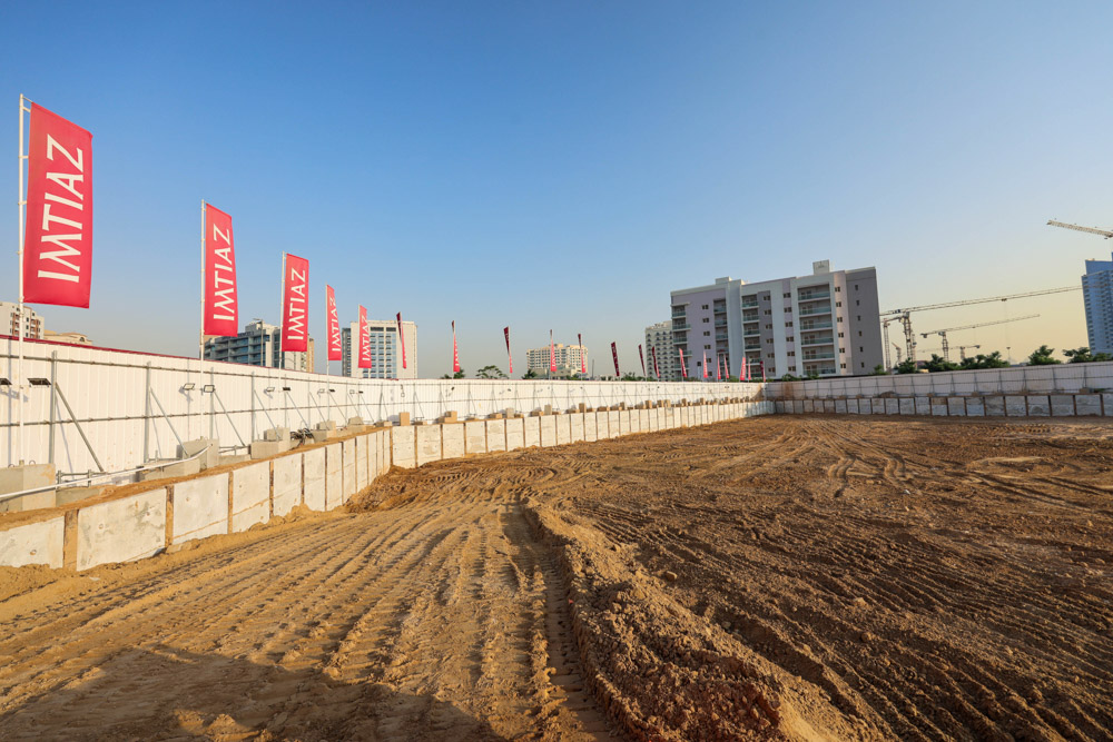 Cove Grand By Imtiaz Construction site with a sandy ground, white barriers, and multiple flags. Buildings are visible in the background.