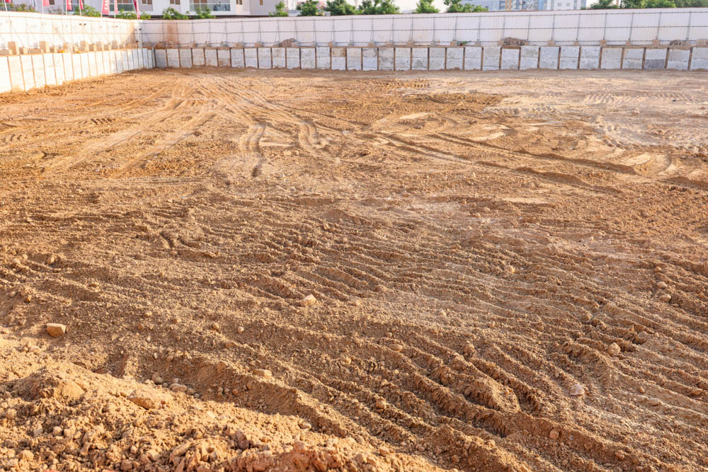 Cove Grand By Imtiaz A large, empty construction site featuring dirt with tire marks and concrete blocks lining the perimeter.