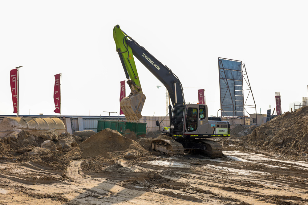 Cotier House 2 By Imtiaz A heavy excavator works on a construction site, digging and moving dirt with banners in the background.