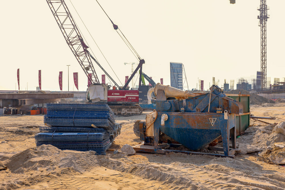 Cotier House 2 By Imtiaz Construction site featuring machinery, a crane, stacked materials, and a sandy landscape under a hazy sky.