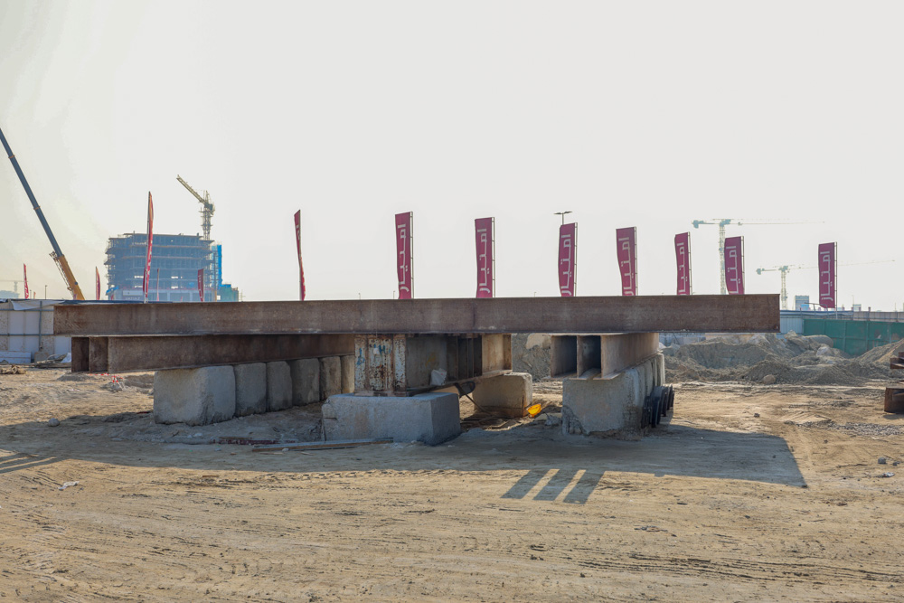 Cotier House 2 By Imtiaz Construction site featuring a large steel beam supported by concrete blocks, with cranes and banners in the background.