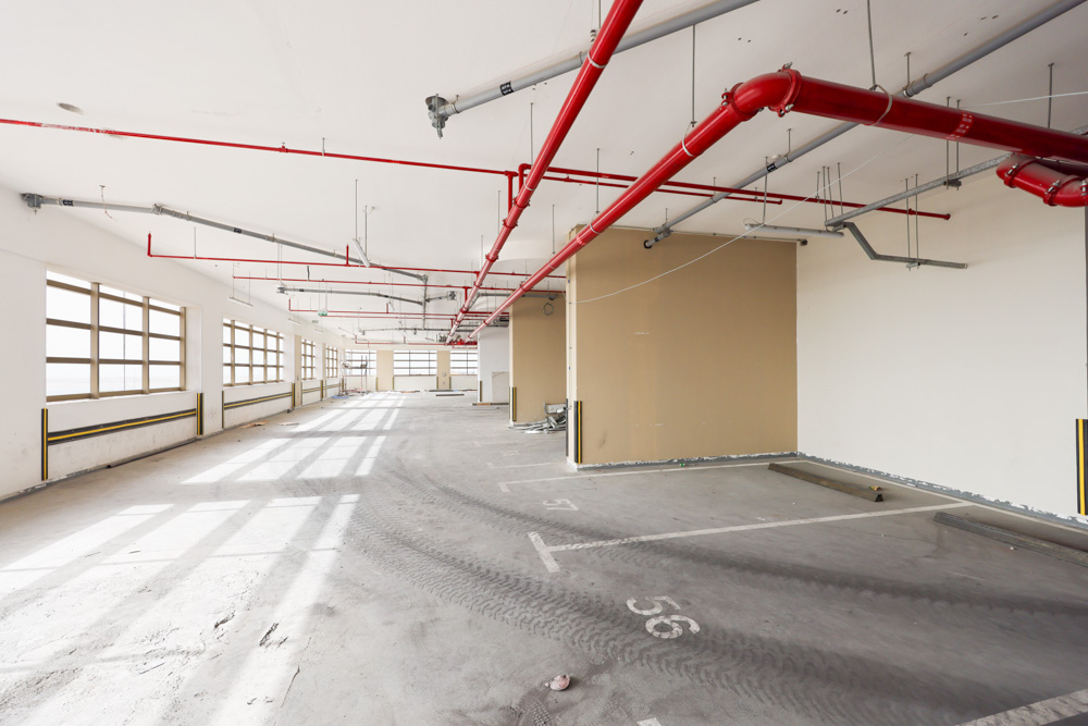 Beach Walk Residence An empty parking garage interior featuring concrete flooring, large windows, and exposed red piping overhead.
