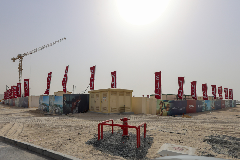 Cotier House 2 By Imtiaz A construction site with red flags, a crane, and advertising banners in a sandy area under a bright sun.