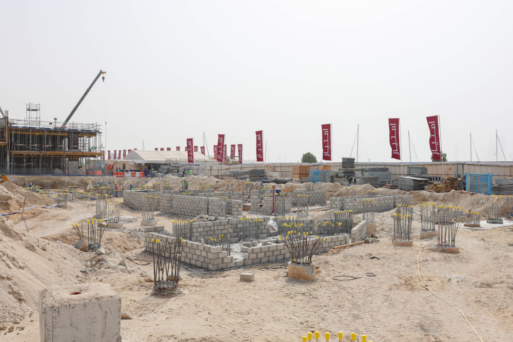 Cotier House 2 By Imtiaz Construction site with concrete blocks and rebar, featuring workers, cranes, and banners in the background.