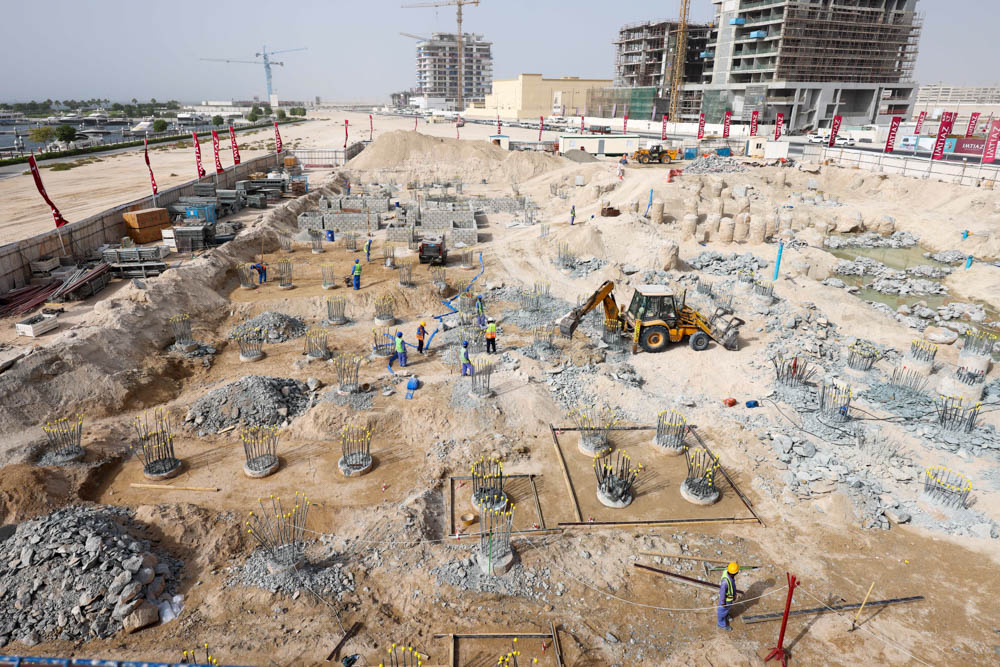 Cotier House 2 By Imtiaz A construction site featuring workers, machinery, and foundations with steel rods amidst piles of gravel and soil.