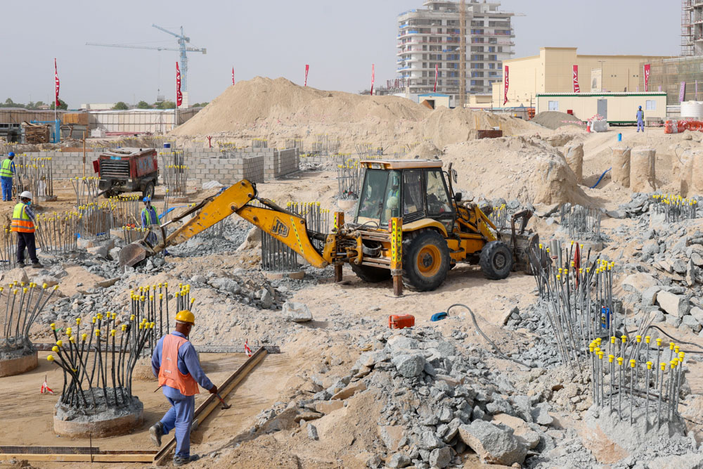 Cotier House 2 By Imtiaz Construction site with workers, machinery, and rebar foundations amidst gravel and sand. Buildings are in the background.