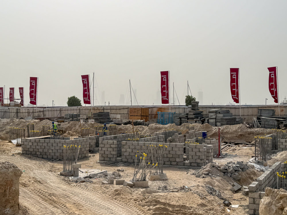 Cotier House 2 By Imtiaz Construction site with stone blocks, workers, and flags in a dusty environment near a waterfront with cranes.