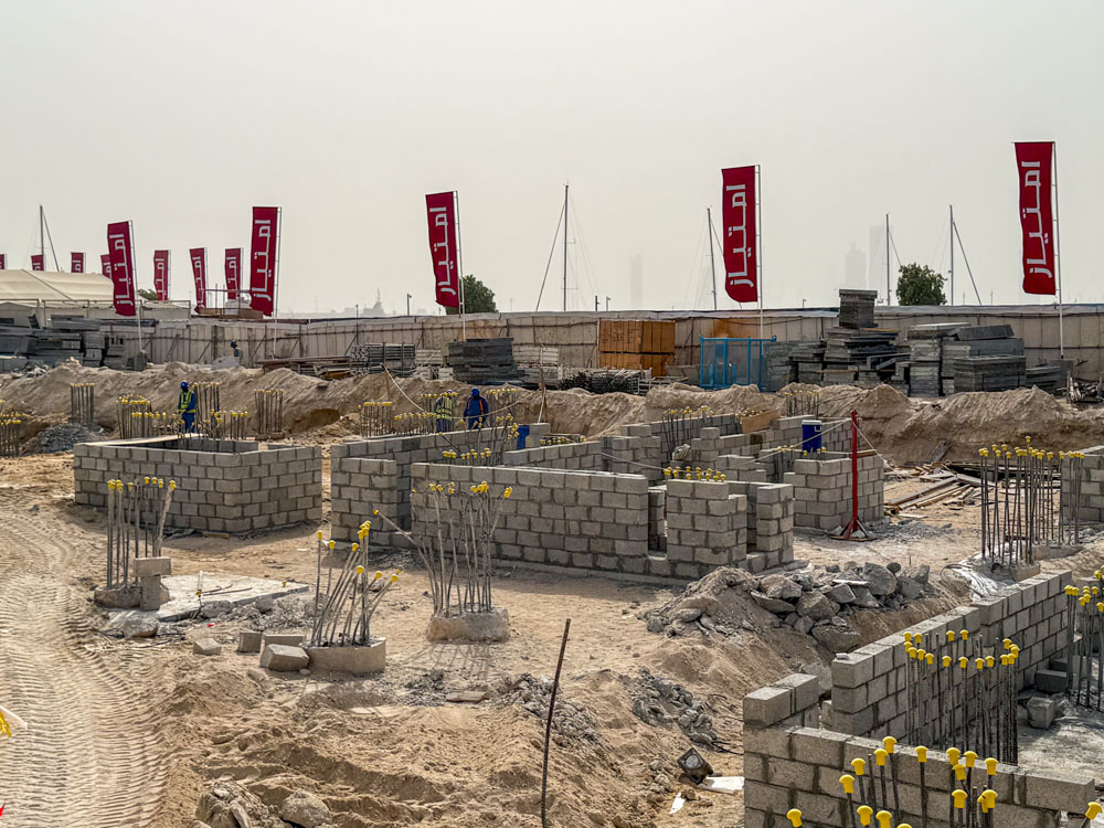 Cotier House 2 By Imtiaz Construction site with concrete blocks, steel reinforcements, and workers under banners in a sandy landscape.