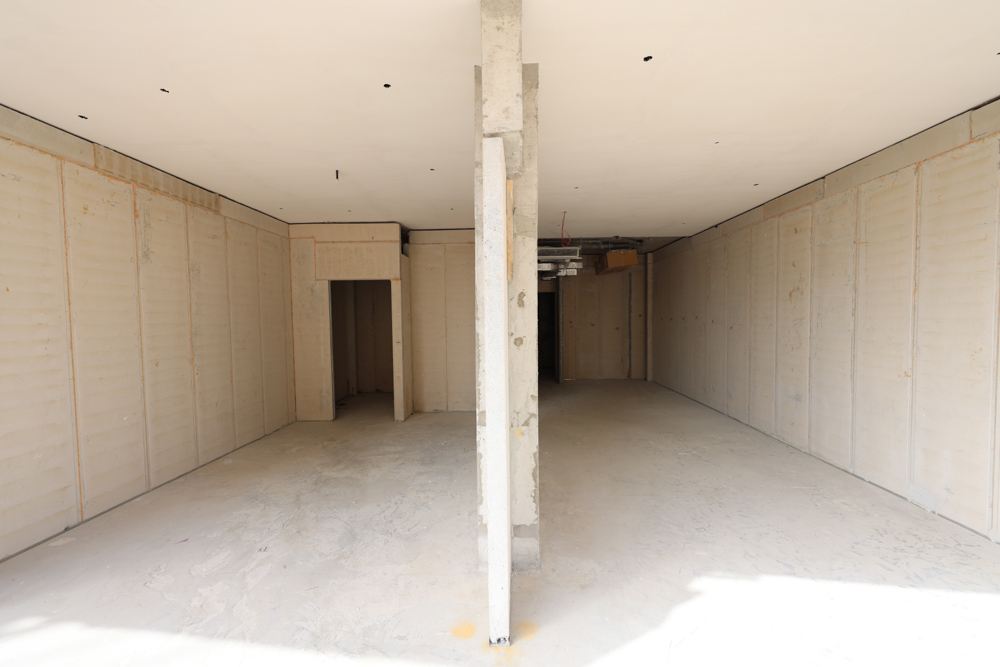 Cove Edition I By Imtiaz 1 A spacious unfinished interior with concrete walls, a central column, and an opening leading to another area.