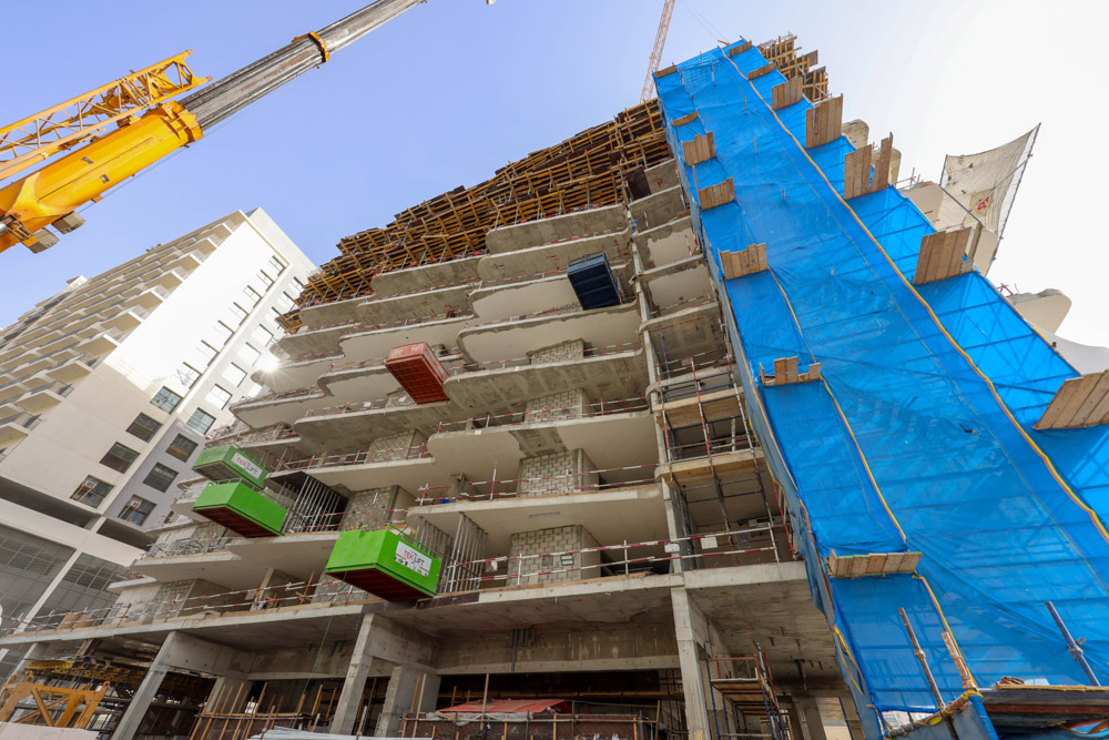 Cove Edition I By Imtiaz 1 A high-rise building under construction with scaffolding, cranes, and protective blue sheeting on its side.