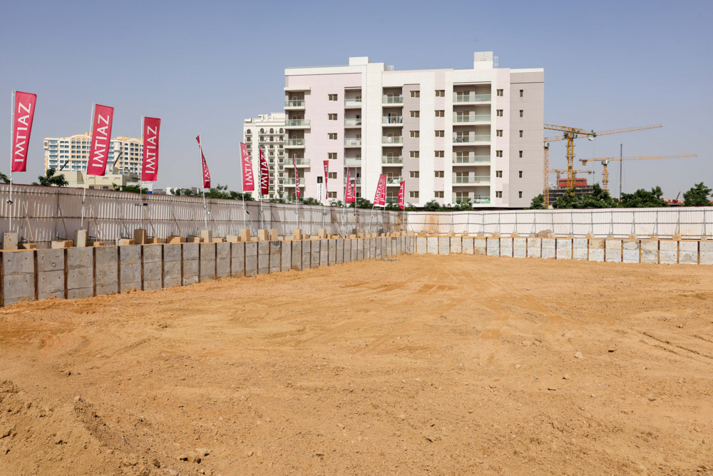 Cove Grand By Imtiaz A construction site with a flat sandy area, concrete walls, and a multi-story building in the background.