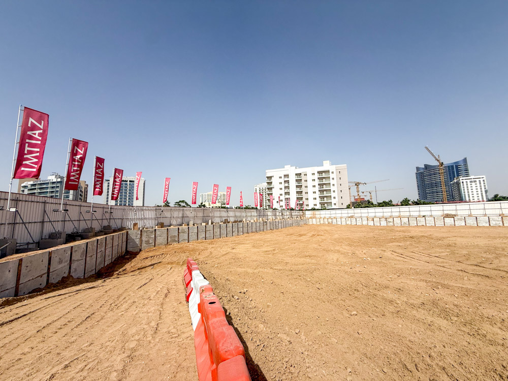 Cove Grand By Imtiaz Construction site with sandy terrain, concrete barriers, and flags; buildings and cranes in the background.