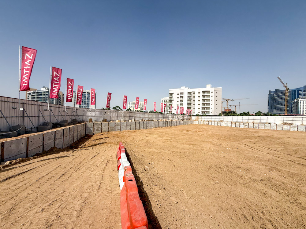 Cove Grand By Imtiaz An empty construction site with sand, concrete barriers, and flags, surrounded by buildings and cranes in the background.