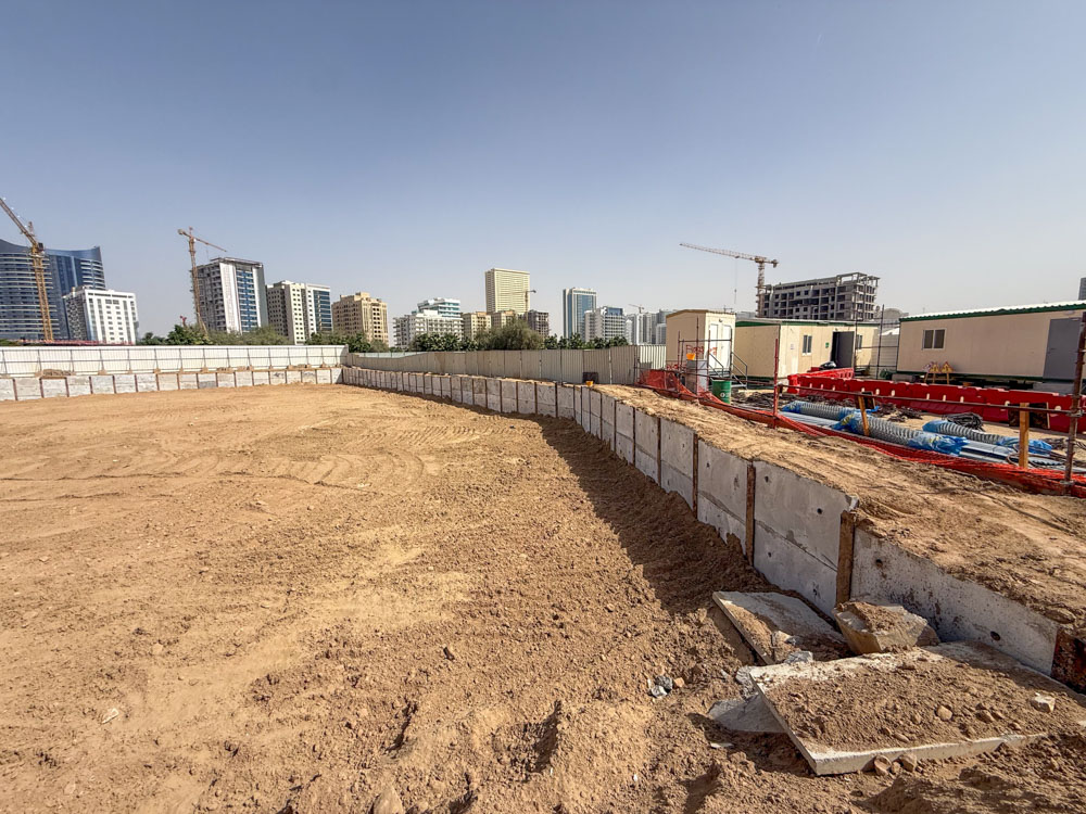 Cove Grand By Imtiaz A construction site features a sandy area bordered by concrete walls, with city buildings visible in the background.