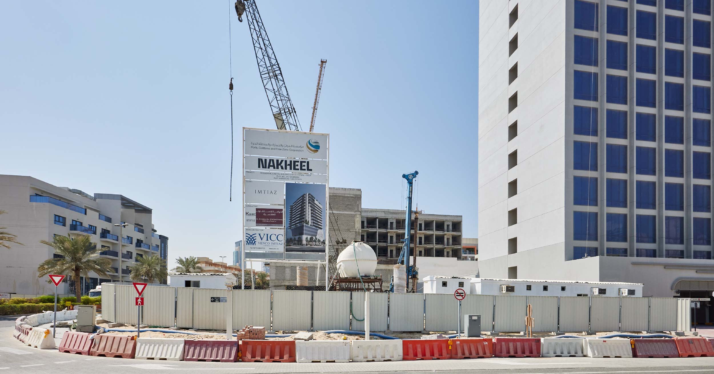 Pearl House By Imtiaz A construction site featuring a large sign, cranes, and surrounding barriers, with nearby modern buildings in view.