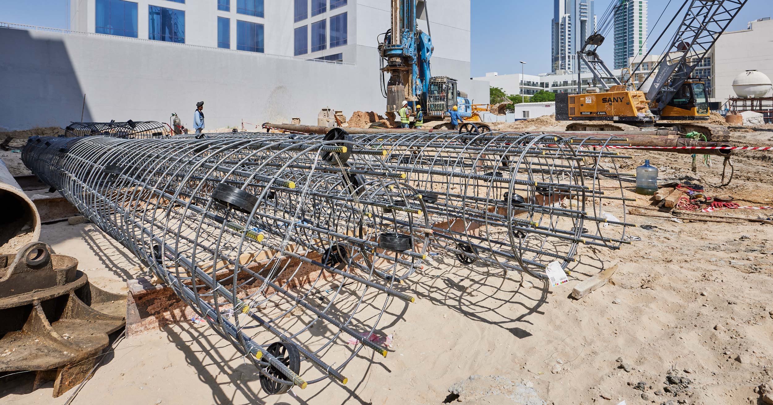 Pearl House By Imtiaz A construction site featuring steel rebar cages on sandy ground, with machinery and workers in the background.