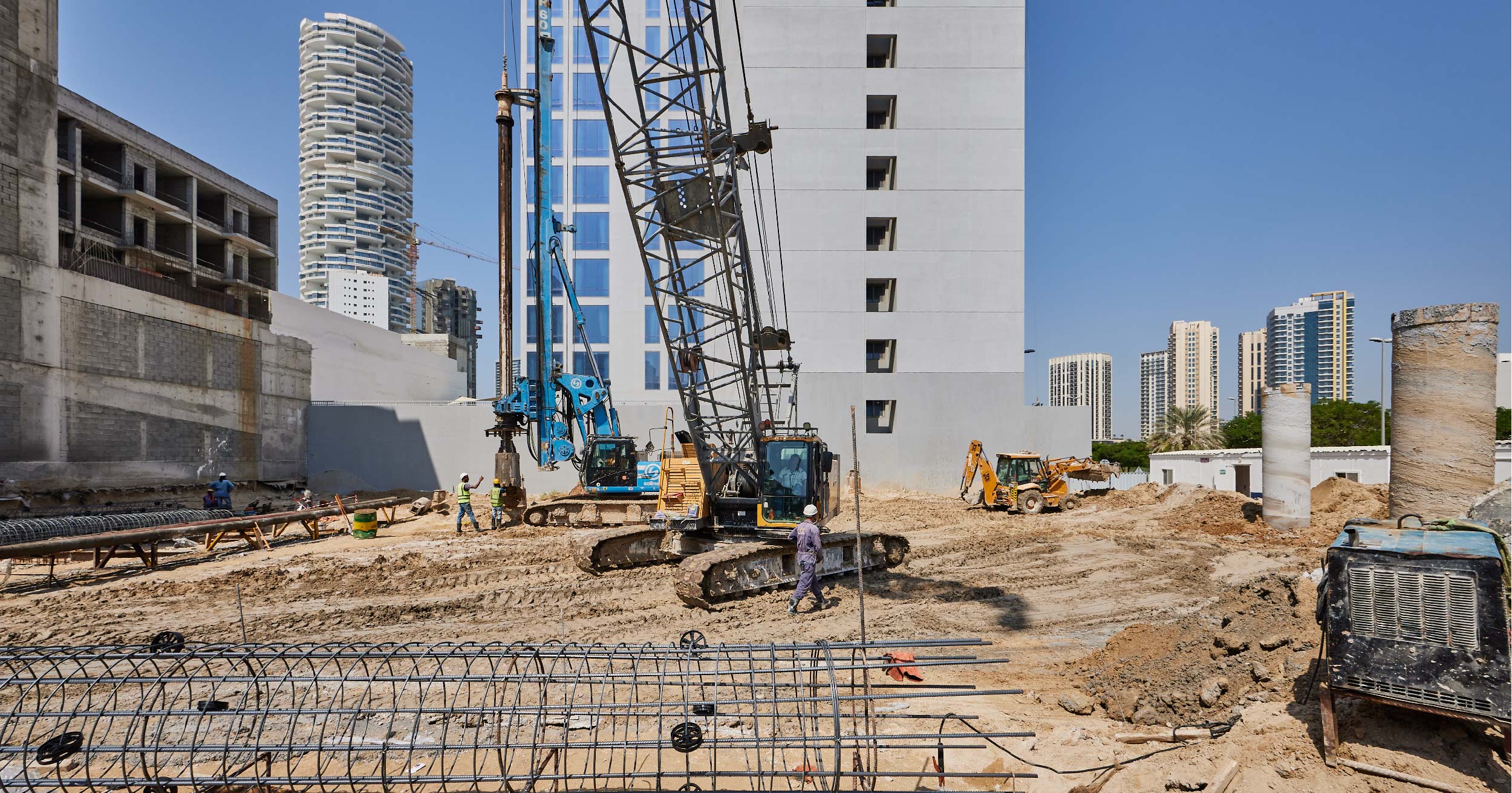 Pearl House By Imtiaz Construction site with heavy machinery, workers, and steel rebar in a sandy area, surrounded by buildings.